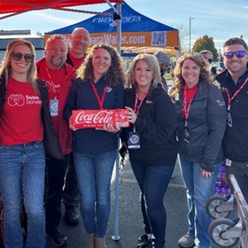 Swire Coca-Cola associates supporting Operation Homefront’s Holiday Meals for Military Program by delivering meals and Coca-Cola beverages. The image shows team members standing together, holding Coca-Cola products, with a tent and blue sky in the background.