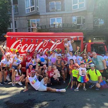 Swire Coca-Cola associates and their families posing together in front of a Coca-Cola truck at the Pride Parade in Seattle, WA. The group is smiling and waving, with many people holding Dasani water bottles to give away to thirsty parade goers.