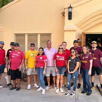 Swire Coca-Cola associates standing in front of a building, supporting the University of Arizona Foundation's Campus Pantry. The image shows team members smiling and posing together, highlighting their efforts in sorting produce and stocking shelves for students and staff facing food insecurity.
